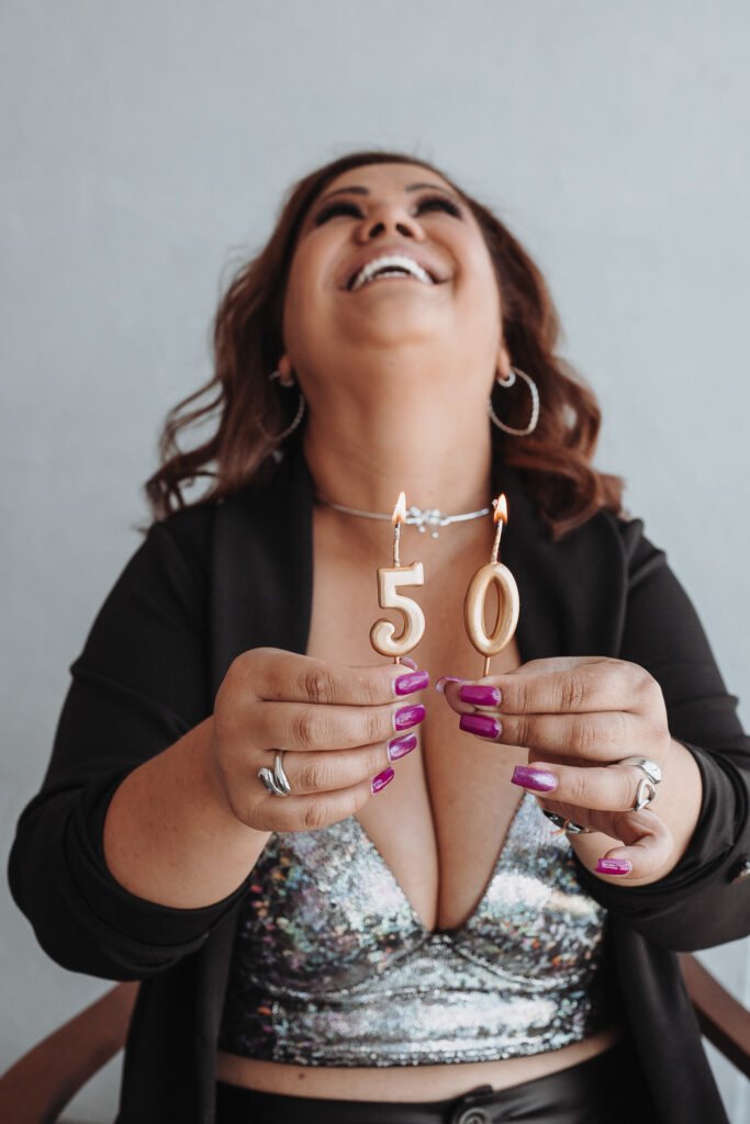 Mulher sorridente segura velas de aniversário douradas com o número '50' acesas. Ela veste um top de lantejoulas prateadas e um blazer preto.